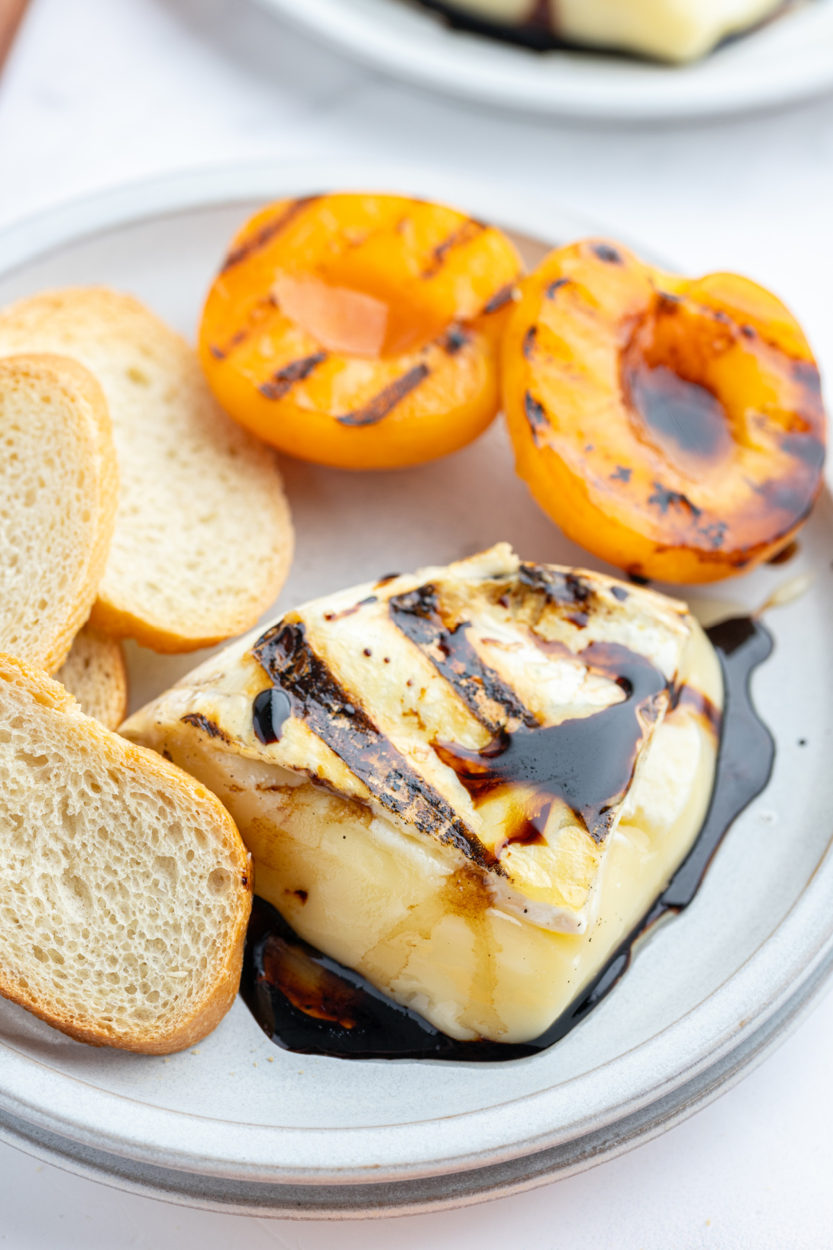 chunk of melted brie on plate with balsamic glaze, baguette and grilled apricots